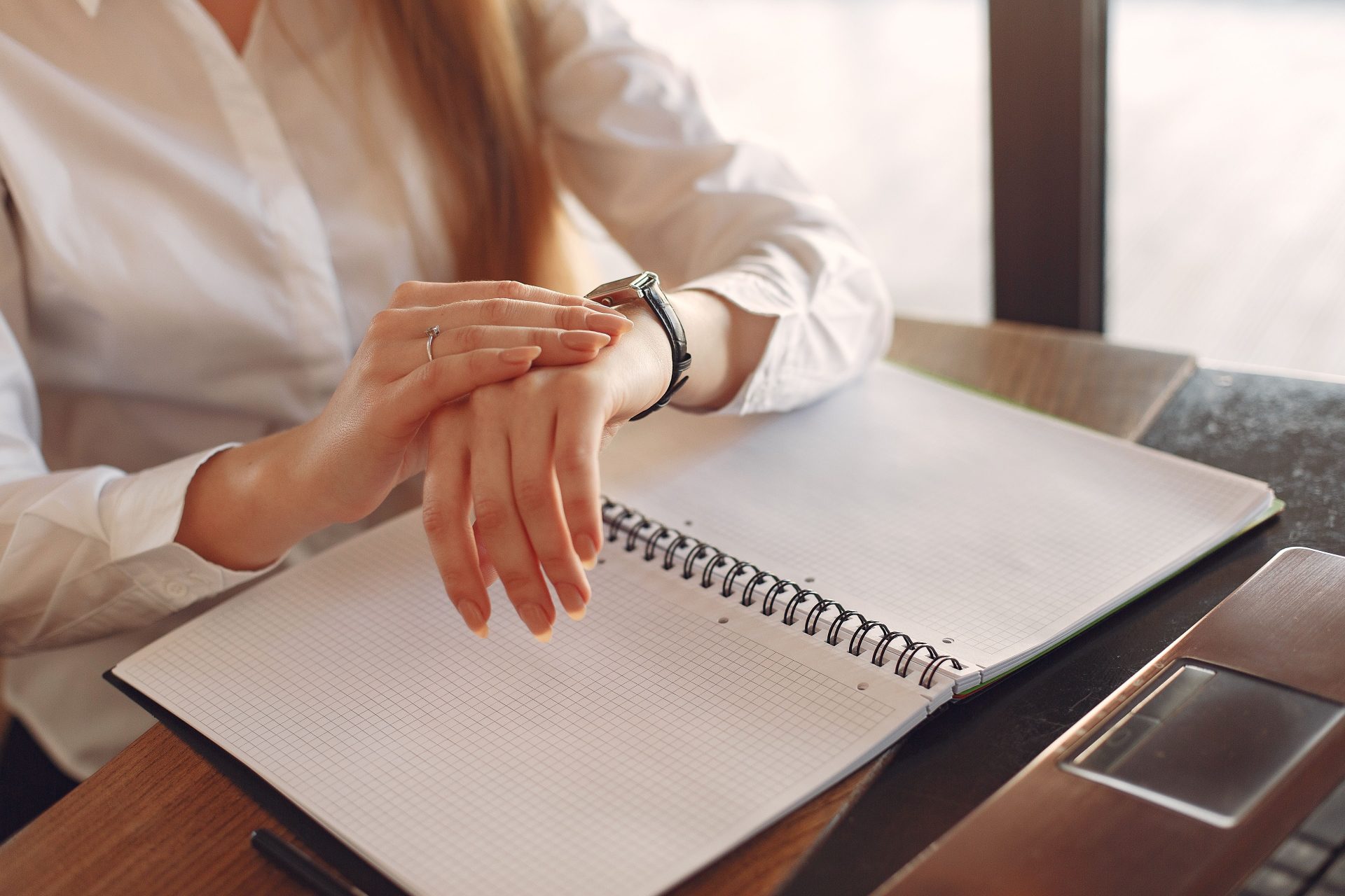 Mujer emprendedora irreconocible vestida formalmente sentada en una mesa de madera cerca de una computadora portátil con una libreta y mirando la hora en un reloj de pulsera.