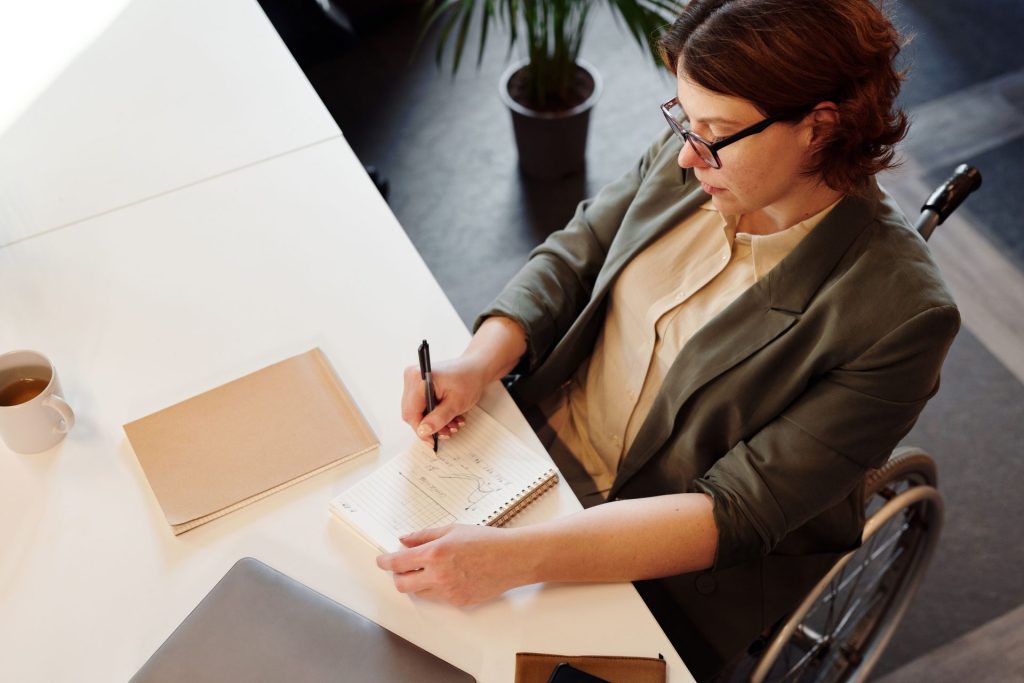 Une femme active en fauteuil roulant prend des notes à un bureau moderne, illustrant la diversité au travail.